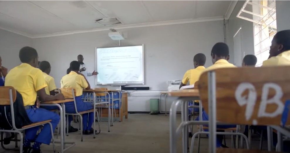 Students at desks in two rows facing a teacher at a smart board screen