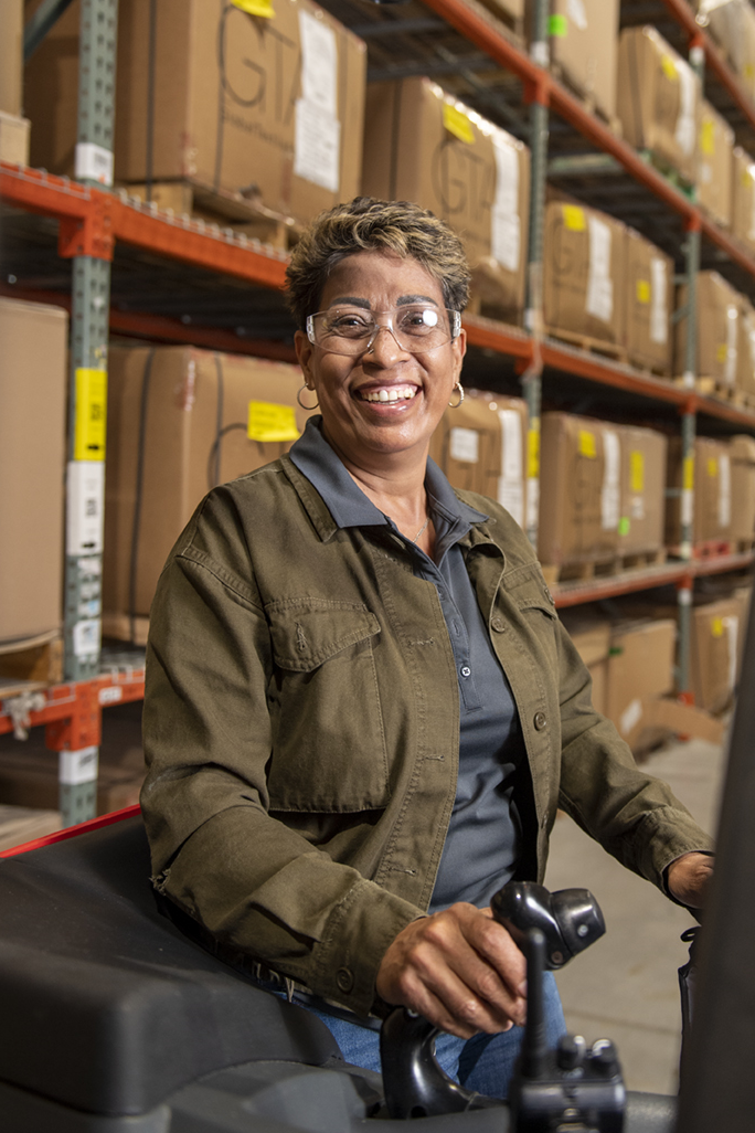 A person wearing safety glasses and a brown jacket smiles while sitting on a forklift inside a warehouse. The background shows rows of stacked cardboard boxes on metal shelves.